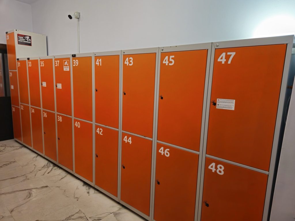 Rząd pomarańczowych szafek z numerami od 28 do 48, umieszczonych w pomieszczeniu wyłożonym marmurową podłogą.