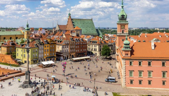 Widok z lotu ptaka na ruchliwy plac miejski z kolorowymi zabytkowymi budynkami, dachami pokrytymi czerwoną dachówką i kościołem z zielonym dachem w pobliżu Warszawiw. Ludzie spacerują i gromadzą się na tętniącym życiem otwartym placu pod częściowo zachmurzonym niebem.