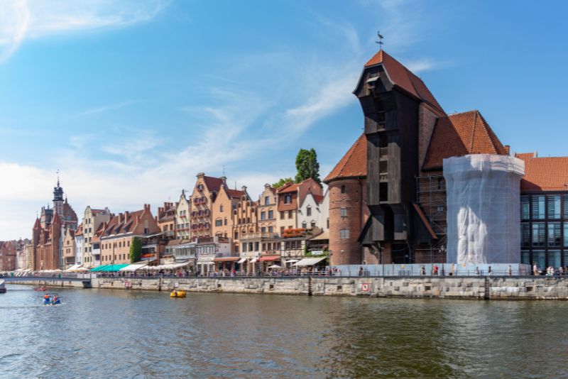 A view of historic buildings in Gdansk along the Motława River, including the characteristic medieval Crane, with surrounding architecture and floating boats.