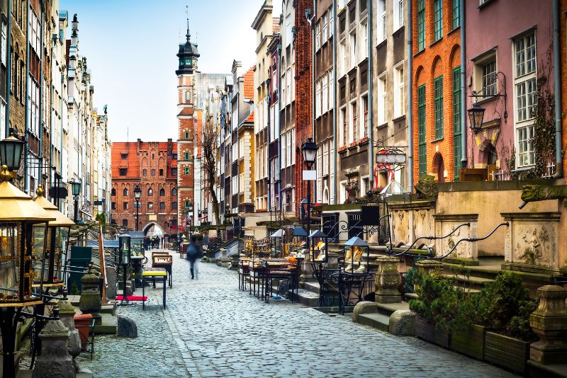 A street in Gdansk with a cobblestone surface, surrounded by historic townhouses with brick facades and decorative elevations, with a visible tower in the distance.
