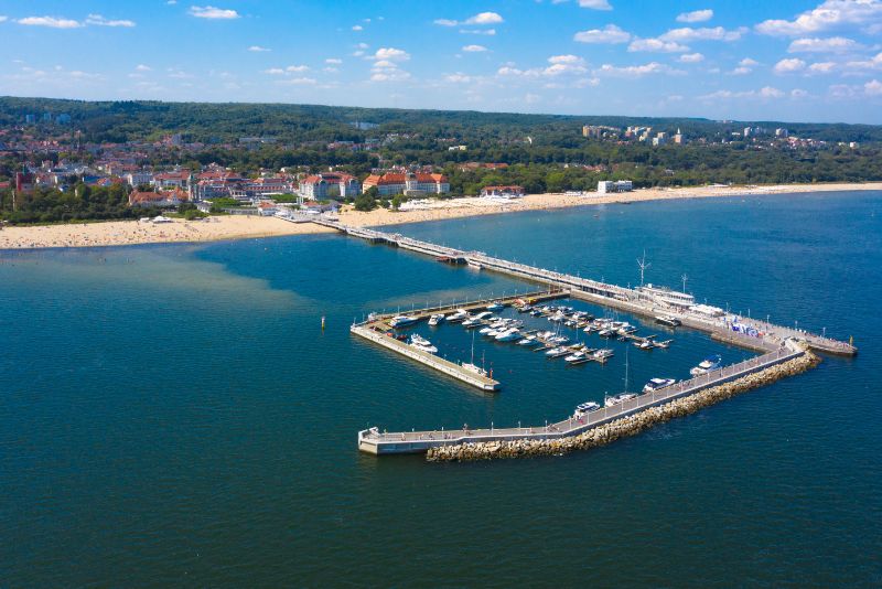 An aerial view of the pier in Sopot with a marina, surrounded by the blue Baltic Sea, with a sandy beach and green forest in the background.