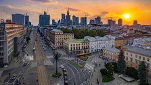 Panorama Warszawy z widokiem na centrum miasta o zachodzie słońca, z ulicą i tramwajem na pierwszym planie.