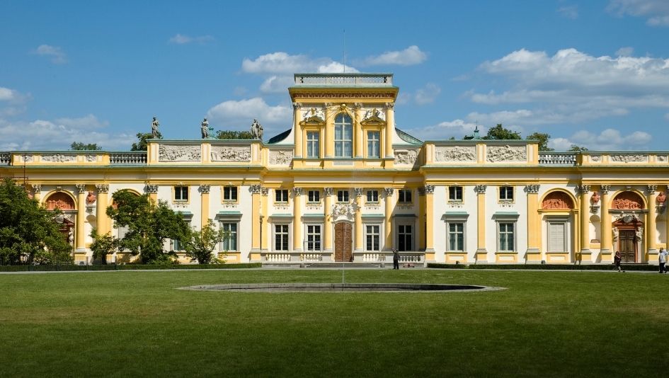 Barokowy pałac z żółtymi i białymi detalami, z dużymi oknami i ozdobnymi elementami, na tle nieba z chmurami.