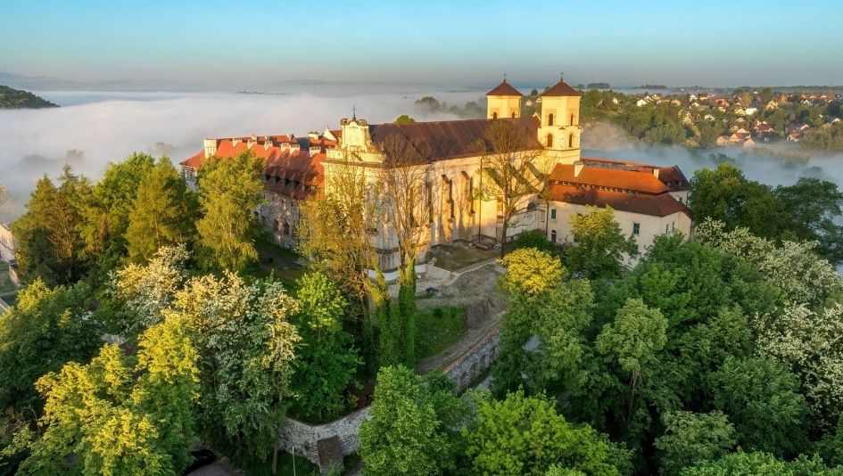 Widok z lotu ptaka na klasztor w Tyńcu usytuowany na wzgórzu, otoczony zielonymi drzewami, z mgłą zalegającą nad okolicą w tle oraz widocznymi budynkami małego miasteczka.