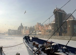 A snow-covered ship moored against the backdrop of Gdańsk's historic buildings, on the Motława River, with the Gdańsk Crane visible and birds flying in the sky.