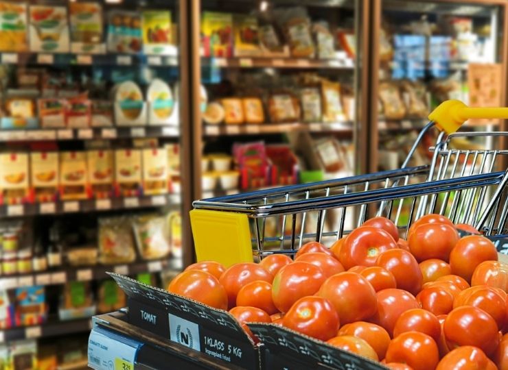 Shopping cart full of red tomatoes with supermarket food product shelves in the background.
