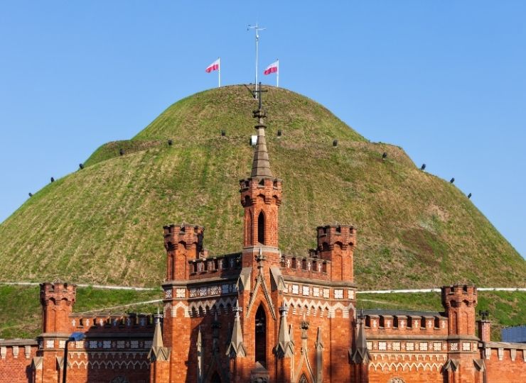Ceglany budynek obronny na tle zielonego kopca z dwiema polskimi flagami na szczycie.