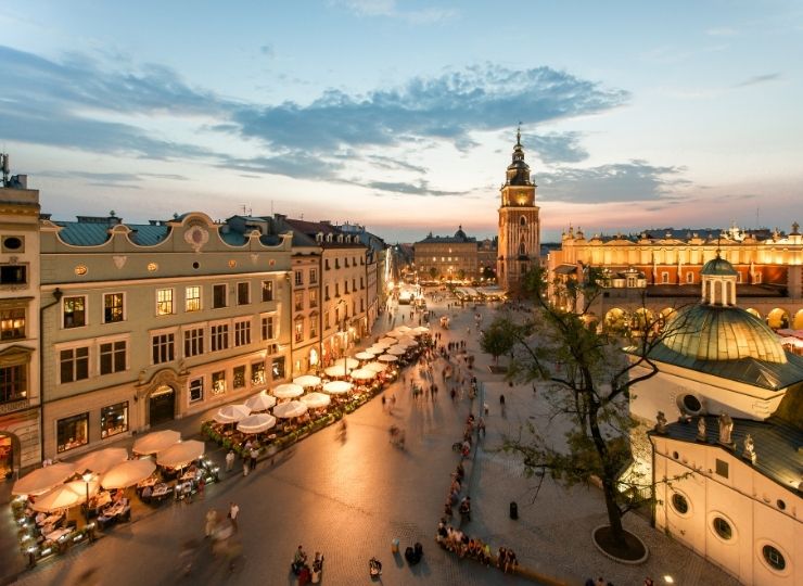Wieczorny widok na Rynek Główny w Krakowie z oświetlonymi budynkami i Sukiennicami, razem z ludźmi spacerującymi i siedzącymi przy kawiarnianych stolikach.