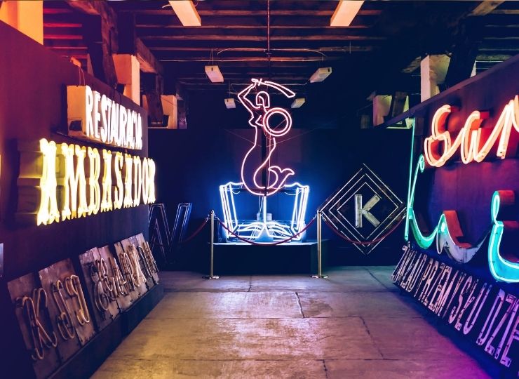 Zabytkowe neony w muzeum, w tym neon przedstawiający postać na szczycie kieliszka oraz napis "Restauracja Ambasador" po lewej stronie.