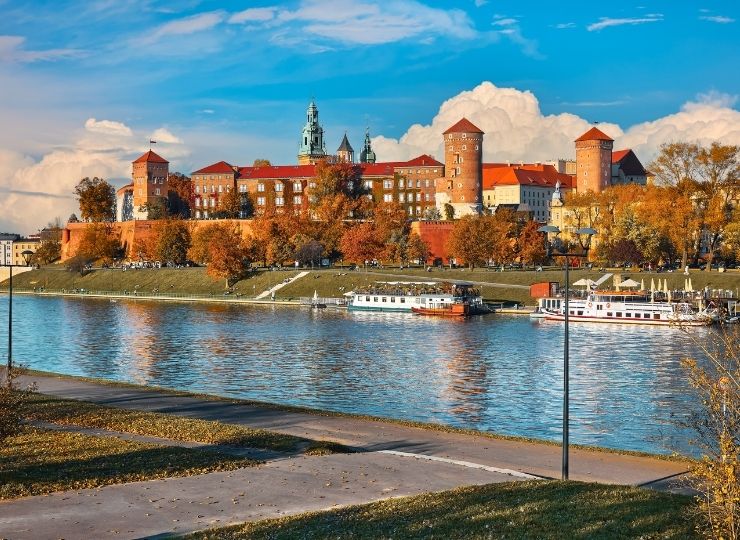 Zamek Królewski na Wawelu w Krakowie widziany z brzegu Wisły, otoczony jesiennymi drzewami, z łodziami zacumowanymi przy nabrzeżu.