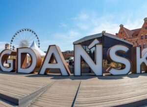 Duże litery „GDAŃSK" stoją na drewnianym podeście, a w tle widoczne są koło widokowe i historyczne ceglane budynki pod bezchmurnym błękitnym niebem.