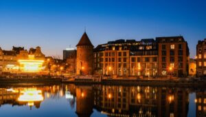 A cityscape of Gdansk – at dusk, with historic and modern buildings along the waterfront. Warm streetlights and building lights reflect on the calm water, and a round stone tower is visible under the clear blue evening sky.