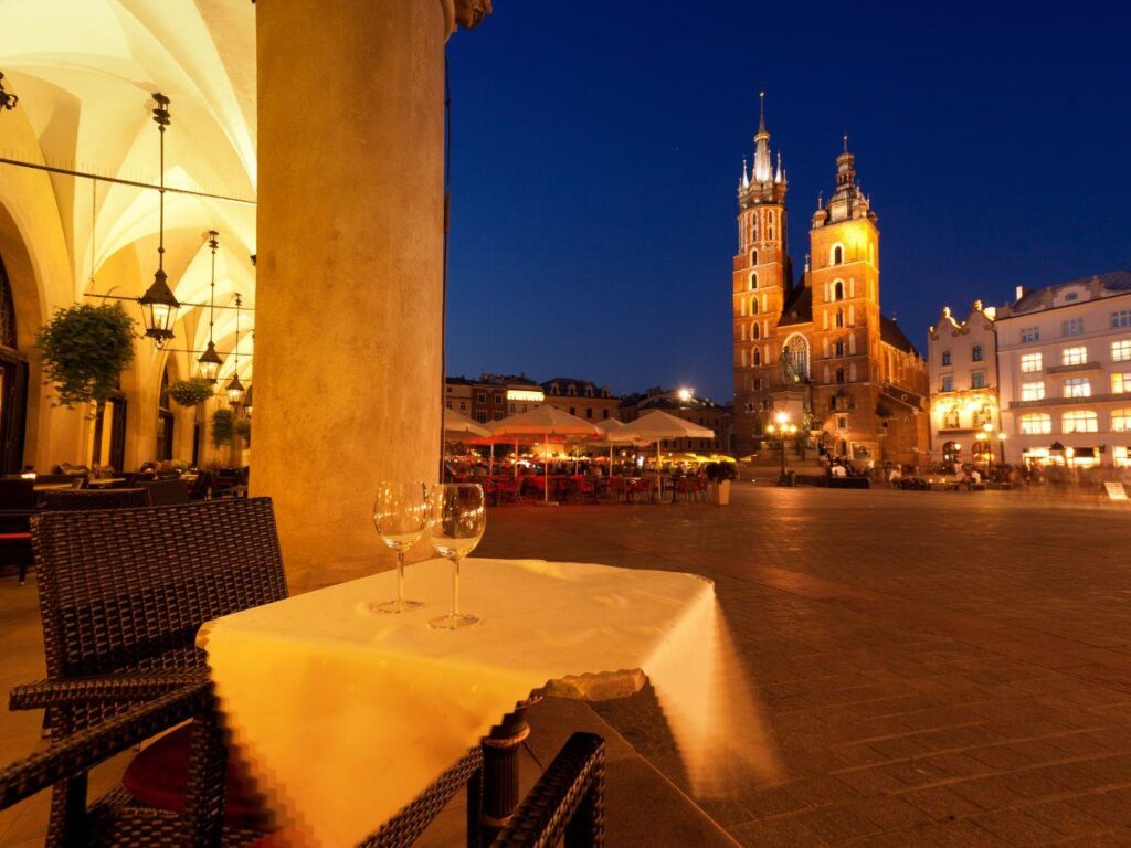 A table with two empty wine glasses at an outdoor café in a European square at dusk, with St Mary’s Basilica and illuminated buildings in the background.