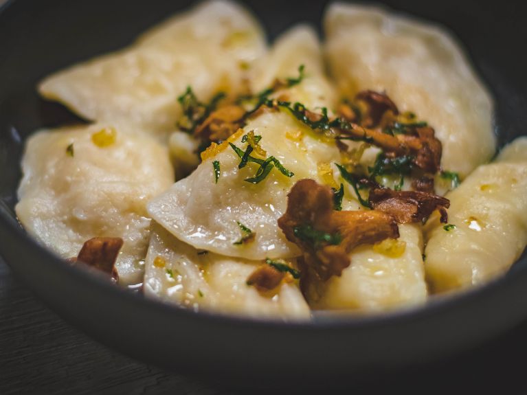A close-up of a bowl filled with dumplings, garnished with chopped herbs and what appears to be sautéed mushrooms, with melted butter or oil glistening on top.