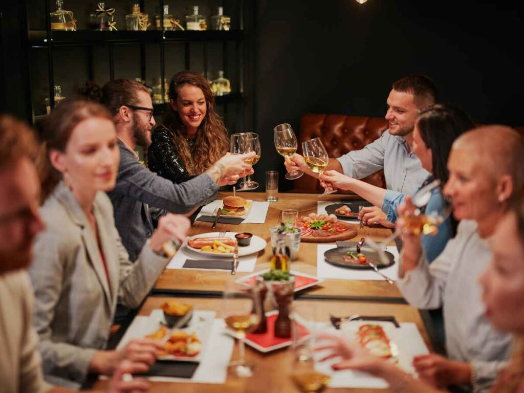 A group of people sitting around a restaurant table, raising glasses in a toast, smiling and enjoying food and drinks in a warm, cosy setting.