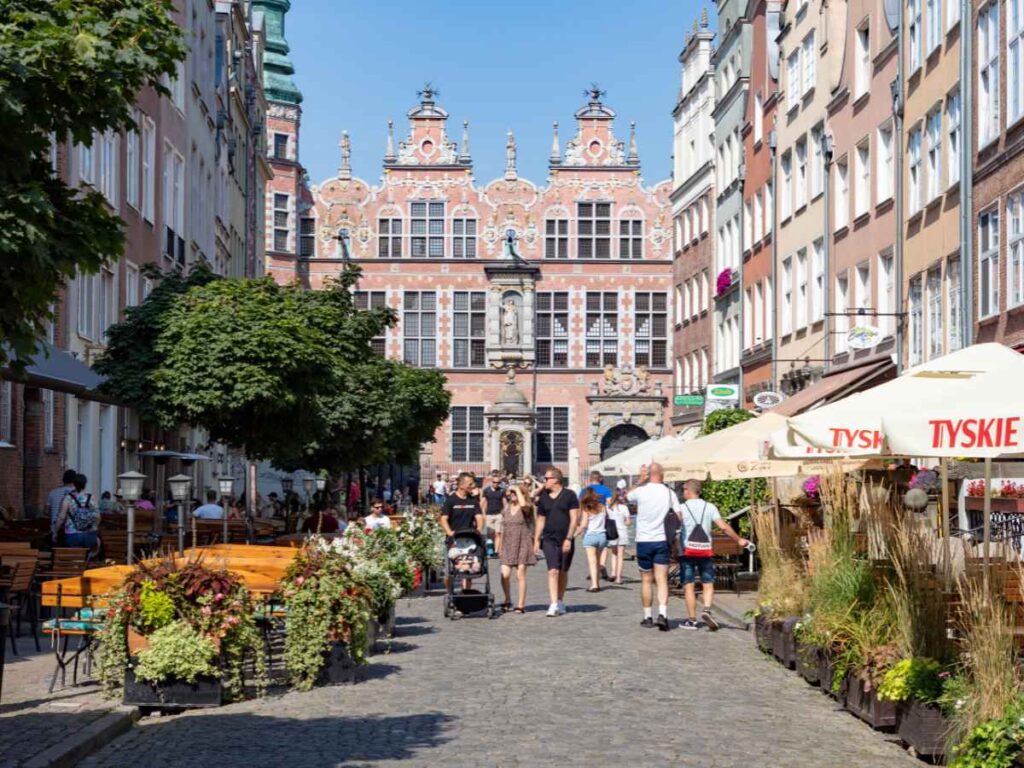 A lively cobbled street lined with outdoor cafés, people walking, potted plants, and historic ornate buildings under a clear blue sky.