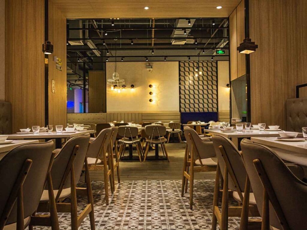 Modern restaurant interior with neatly arranged tables and chairs, warm lighting, wooden accents, tiled floor, and decorative wall elements, creating a cosy and inviting dining atmosphere.