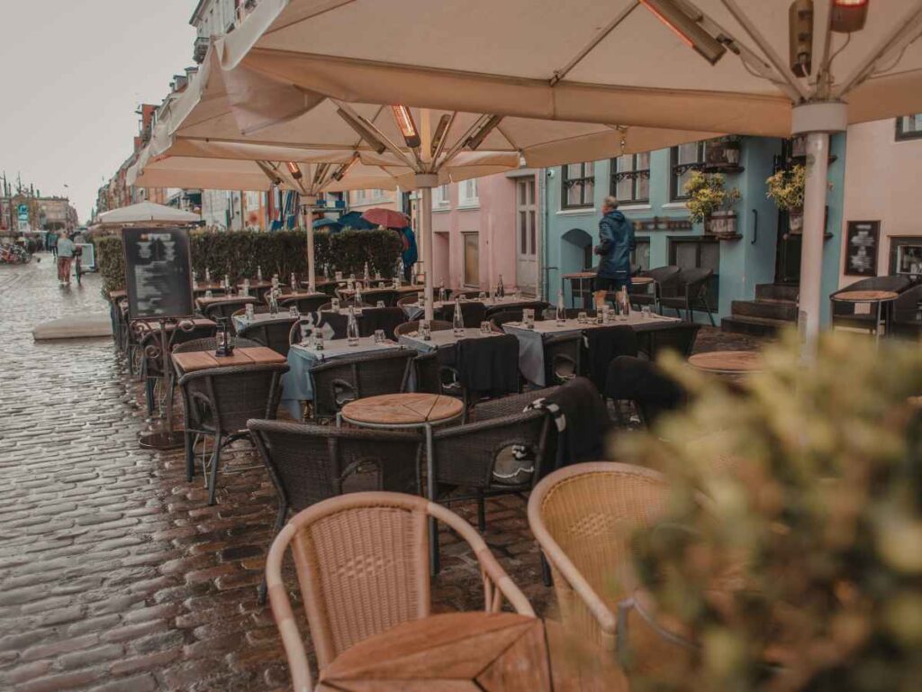 Outdoor café with empty tables and chairs on a wet cobblestone street under large umbrellas. A person is walking in the background, and the atmosphere appears to be cool and rainy.